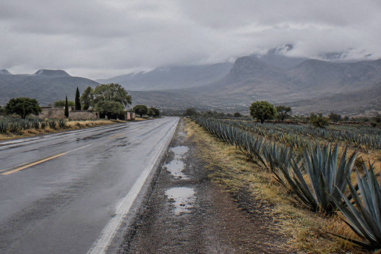 Empty two-lane highway crossing the dry Mezquital Valley toward distant hazy mountains.