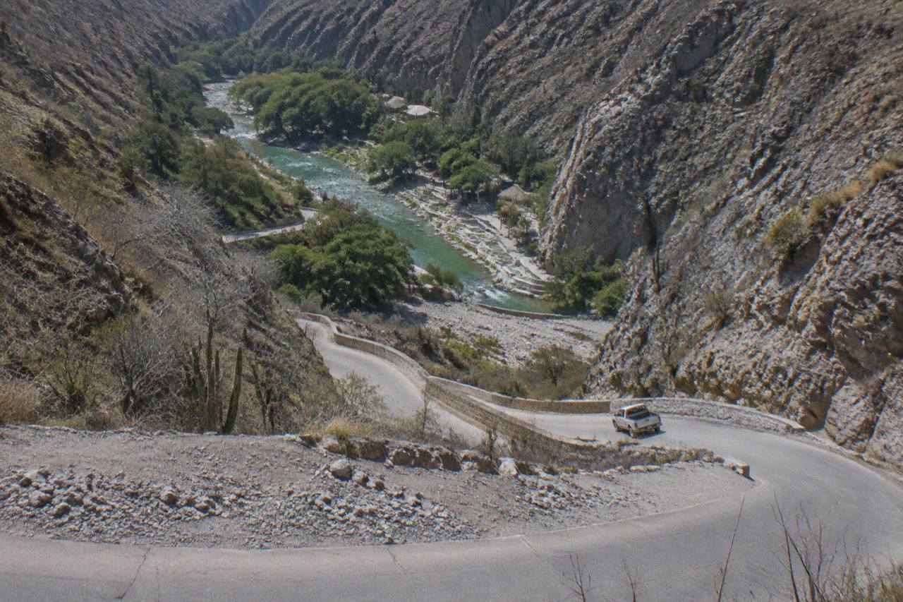 A paved mountain road with tight curves descending between dry canyon walls toward green vegetation below