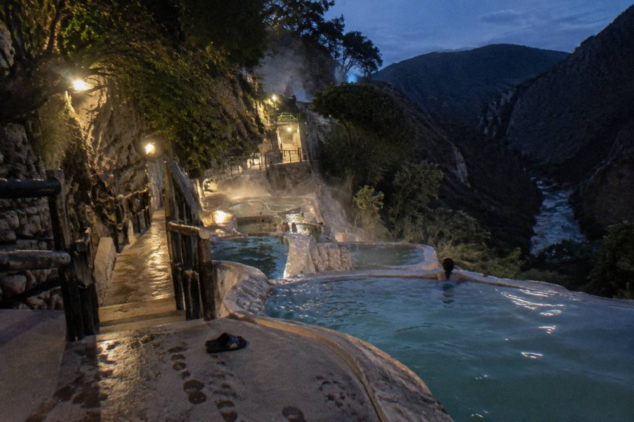 Stepped natural thermal pools built into a canyon cliffside with steam rising from turquoise water