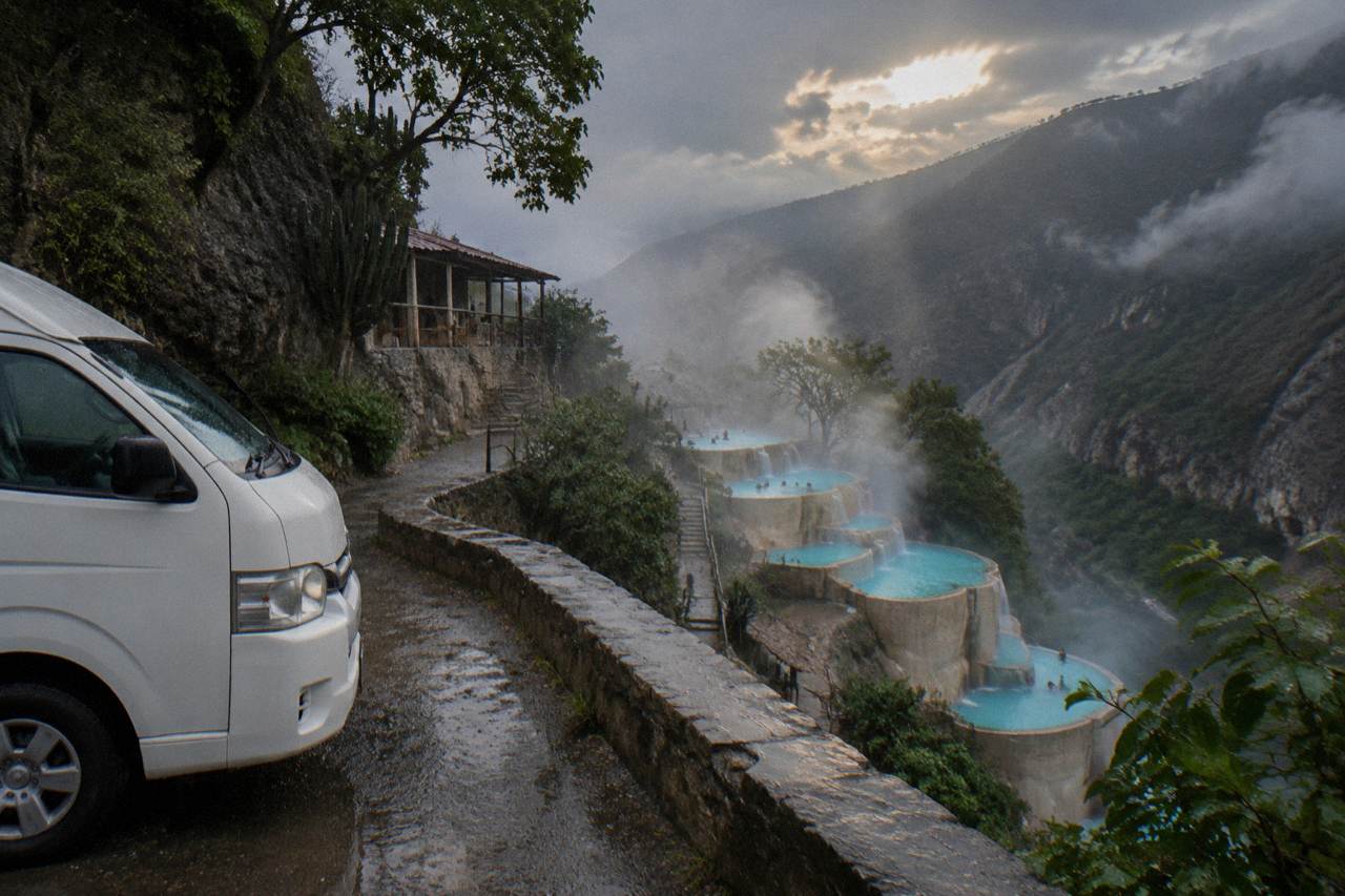 Tour van parked on a canyon overlook above steaming turquoise pools in the valley below