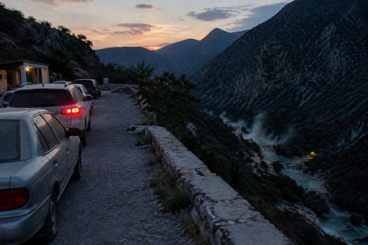 Vehicles parked at a canyon rim lot with steam and turquoise water visible in the valley far below