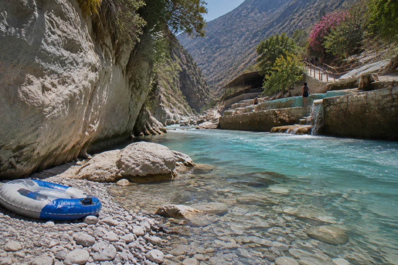 A shallow turquoise thermal river flowing between rock walls below the pools