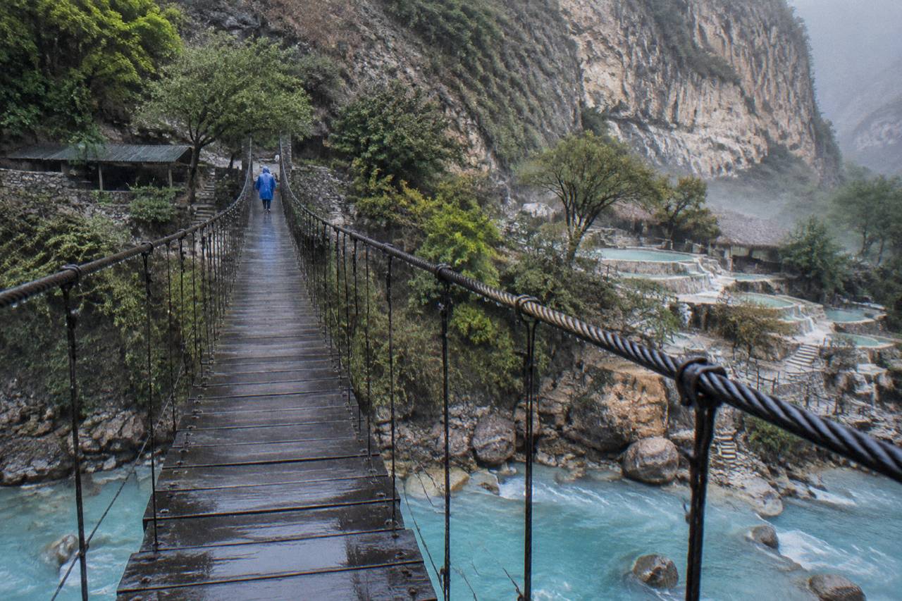 A narrow hanging bridge crossing a green canyon with pools visible on the far wall
