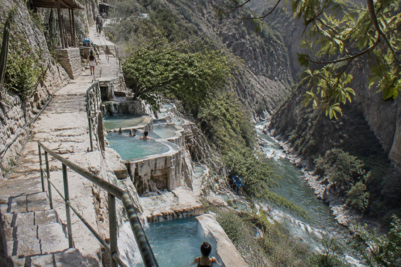 Steep green canyon walls rising above a turquoise thermal river with pools along the cliffside